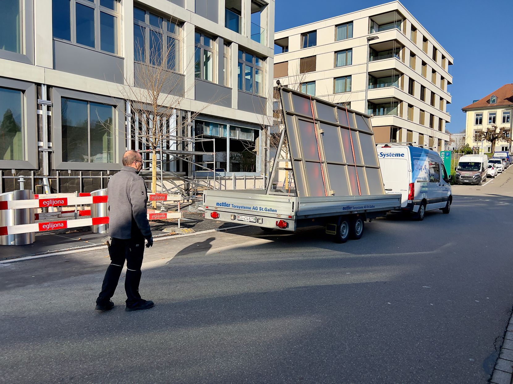 Ein Handwerker steht auf der Strasse und hilft seinem Kollegen einen Lieferwagen mit Anhänger zu manövrieren. Im Hintergrund sind zwei mehrstöckige Neubauten zu sehen.